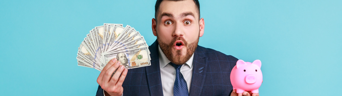 Surprised businessman wearing official style suit looking at camera with shocking expression, holding piggy bank and dollar banknotes in hands. Indoor studio shot isolated on blue background.