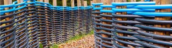 Autumn path with fallen leaves between a fence made of twisted plastic pipes in black and blue braided between wooden posts. Alternative use of polypropylene plumbing pipes in landscape design.