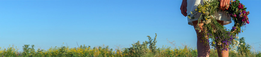 A girl in a white dress holds a wreath of wild flowers. She stands barefoot on green grass against a blue sky.